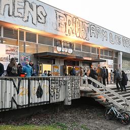 Ein flaches Gebäude mit Graffiti wird von untergehender Sonne beschienen, Menschen stehen auf der umlaufenden Veranda. 