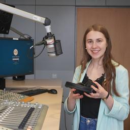 Eine junge Frau steht an einem Mikro und lächelt in die Kamera. 