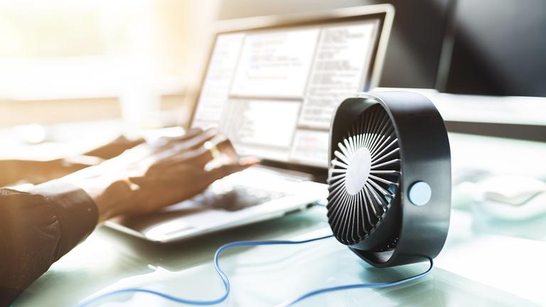 Eine Frau schreibt am Laptop und neben ihr steht ein kleiner Ventilator auf dem Schreibtisch.