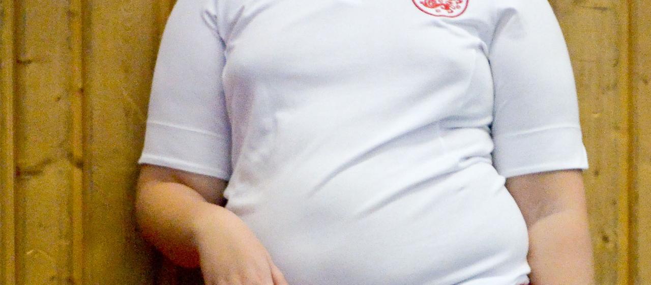 Ein Jugendlicher mit Übergewicht steht an der Wand einer Turnhalle und trägt ein Fußball-Trikot.