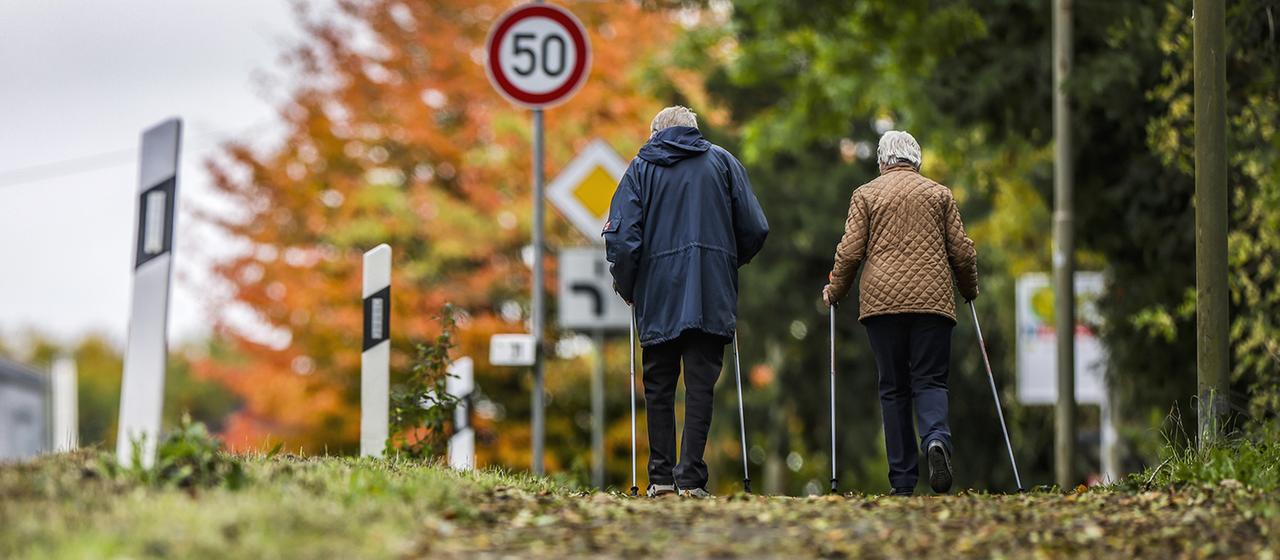 Zwei ältere Personen gehen im Herbst mit Wanderstöcken auf einem Gehweg an einer Straße entlang.