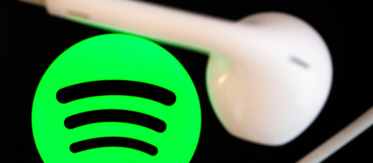  Kopfhörer liegen auf dem Bildschirm eines Smartphones, auf dem das Logo vom Musikstreaming-Dienst Spotify angezeigt wird.