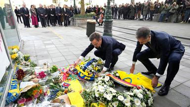 Zwei Männer legen vor einer in Distanz stehenden Menge Kränze nieder.