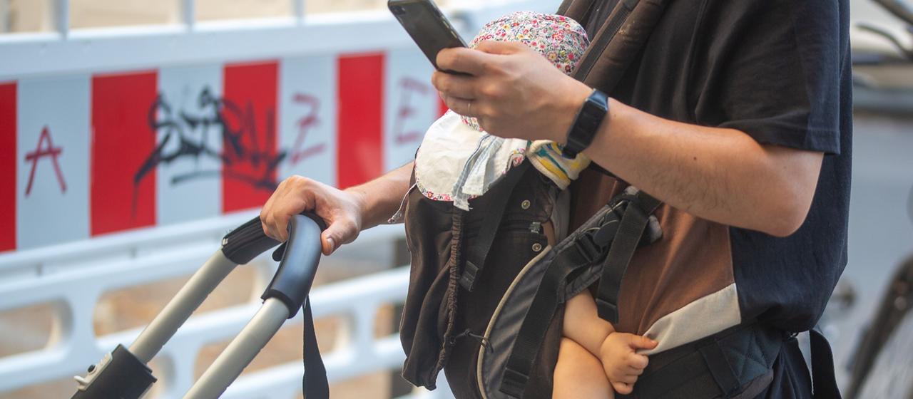 Ein Mann schiebt einen Kinderwagen und trägt gleichzeitig ein Baby in einer Tragehilfe. Dabei schaut er auf sein Smartphone.