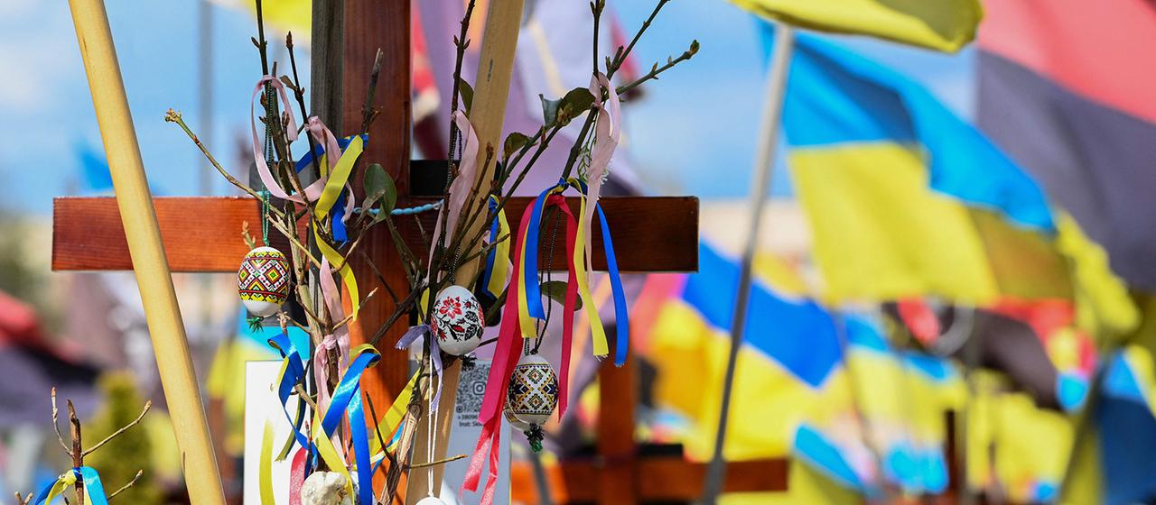 Hinter einem mit Ostereiern geschmückten Kreuz sind ukrainische Flaggen zu sehen.