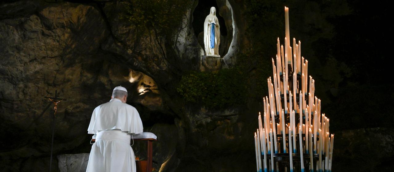 Papst Leo XIV. kniet in der Grotte von Lourdes neben einer Pyramide aus brennenden Kerzen vor der Statue der Heiligen Jungfrau Maria.