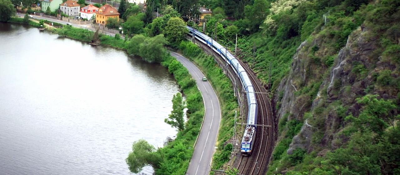 Eine Eisenbahn fährt durch ein Tal an einem Gewässer entlang.