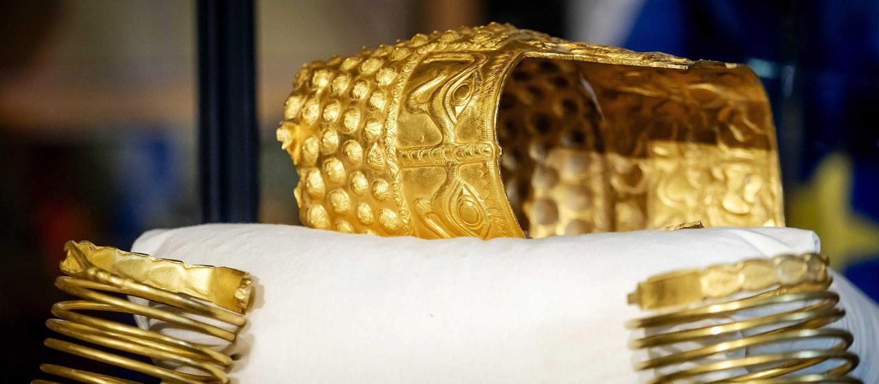 Zwei goldene Armreifen und ein verzierter goldener Helm liegen auf einem weißen Kissen.
