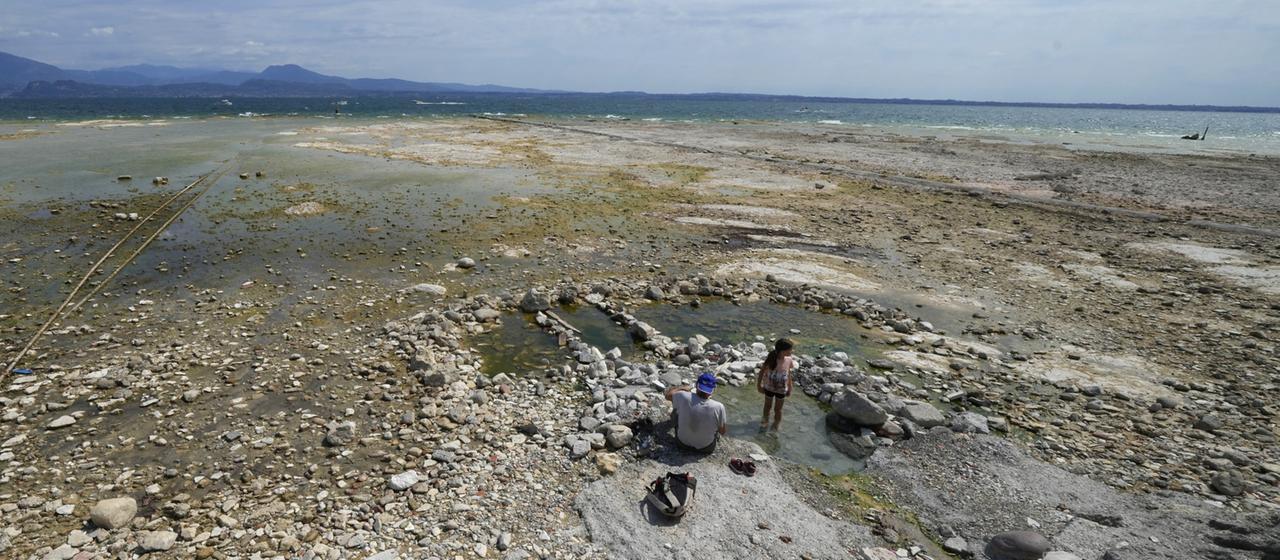 Ein Mann sitzt neben einem Kind am Gardasee. Der Wasserstand des Gardasees war nach einer schweren Dürre stark gesunken, wodurch die Steine, die die Halbinsel von Sirmione umgeben, freigelegt wurden.  | dpa Ein Mann sitzt neben einem Kind am Gardasee. Der Wasserstand des Gardasees war nach einer schweren Dürre stark gesunken, wodurch die Steine, die die Halbinsel von Sirmione umgeben, freigelegt wurden.
