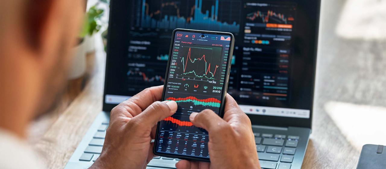 Eine Person überprüft Börsenkurse auf einem Smartphone- und Laptopbildschirm.