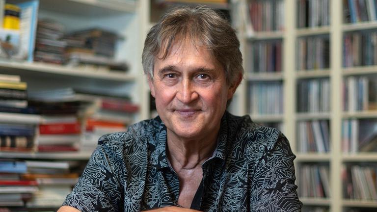 Matthias Creutziger: Ein Mann in gemusterten Hemd sitzt rittlings auf einem Stuhl vor Buch- und CD-Regalen.