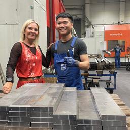 Katrin Fischer und Vin stehen in der Halle der Schmalkaldener Maschinenmesser GmbH. Vor ihnen auf einem Tisch liegen gestapelte Metallteile.