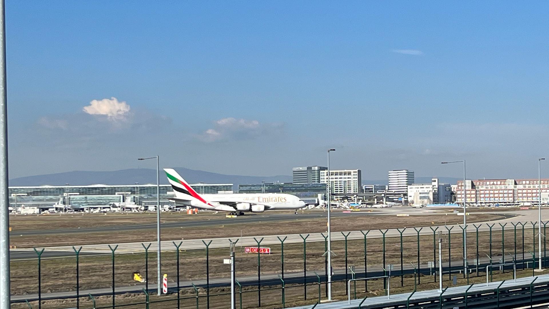 Ein A380-Flugzeug der Fluggesellschaft Emirates auf einem Rollfeld des Flughafens Frankfurt, fotografiert Ã¼ber einen Zaun hinweg. Der Flieger ist weiÃ mit darauf lackierten roten Streifen. | hr
