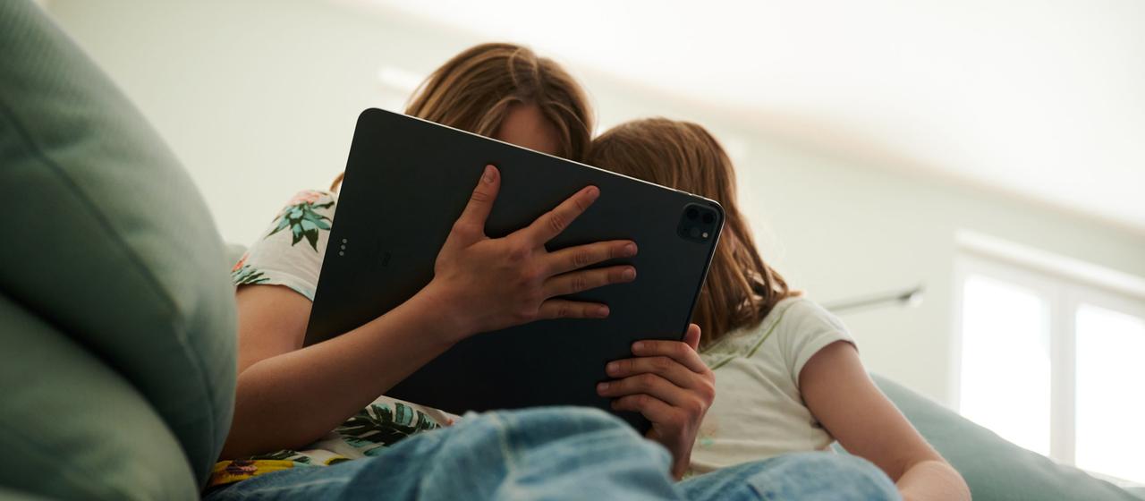 Zwei Schwestern sitzen auf der Couch und gucken in das Tablet ihrer Eltern.