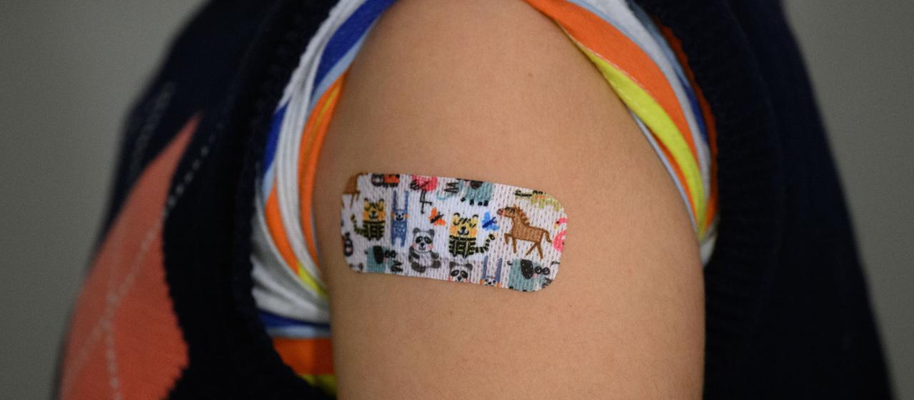 Ein Pflaster mit bunten Tiermotiven klebt nach einer Impfung auf dem Oberarm eines Kindes.