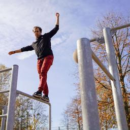 Ein Mann balanciert mit ausgestreckten Armen auf einer Metallstange im Freien. Weitere Stangen sind im Vordergrund.