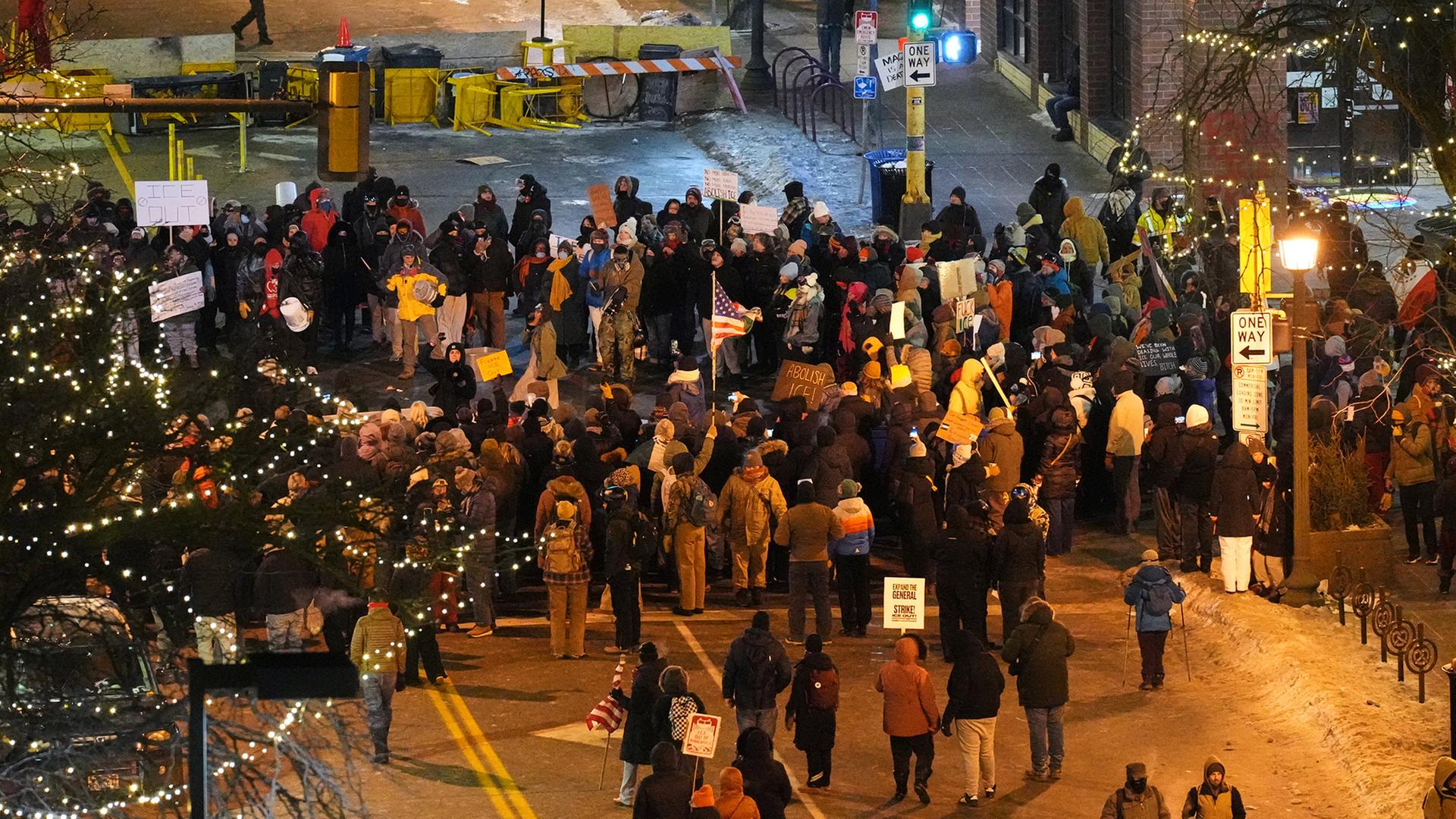 Eine große Gruppe Menschen demonstriert bei winterlichen Temperaturen auf einer beleuchteten Straße, einige halten Plakate und Schilder, darunter eine amerikanische Flagge. | AP