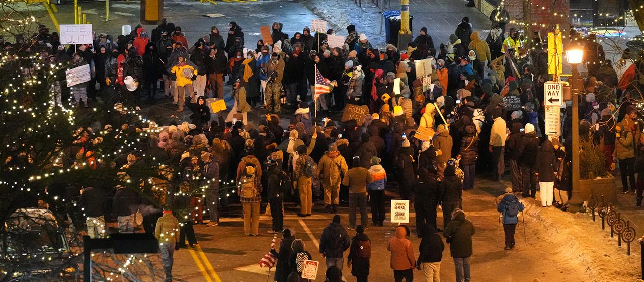 Eine große Gruppe Menschen demonstriert bei winterlichen Temperaturen auf einer beleuchteten Straße, einige halten Plakate und Schilder, darunter eine amerikanische Flagge.