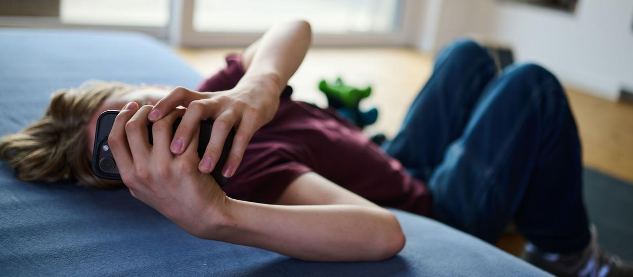 Ein Teenager guckt auf ein Handy, während er auf Boden und Matratze liegt.