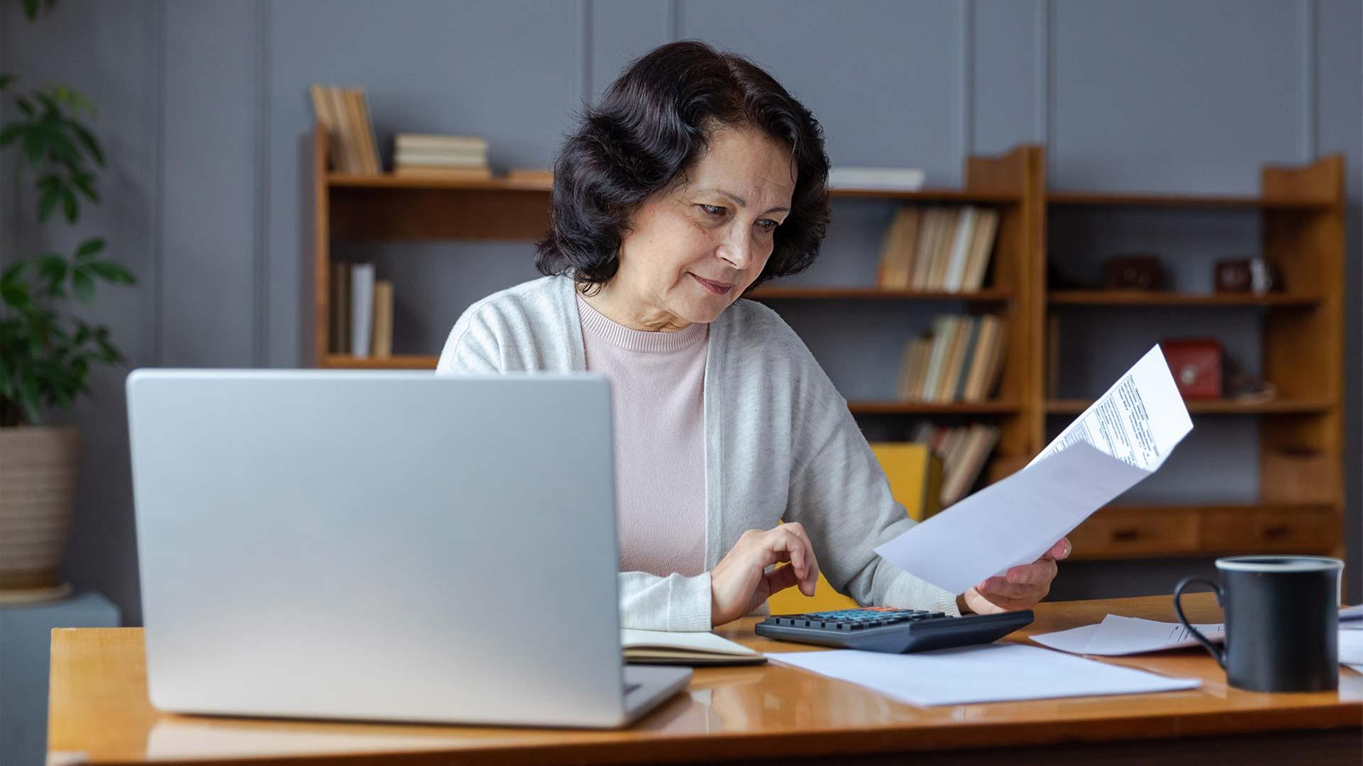 Eine ältere Frau sitzt an einem Schreibtisch mit Laptop und Kalkulator. | picture alliance / Zoonar