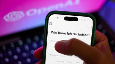 Ein Smartphone mit dem KI-Chatbot ChatGPT ist vor dem Logo des US-Softwareunternehmens OpenAI zu sehen. 