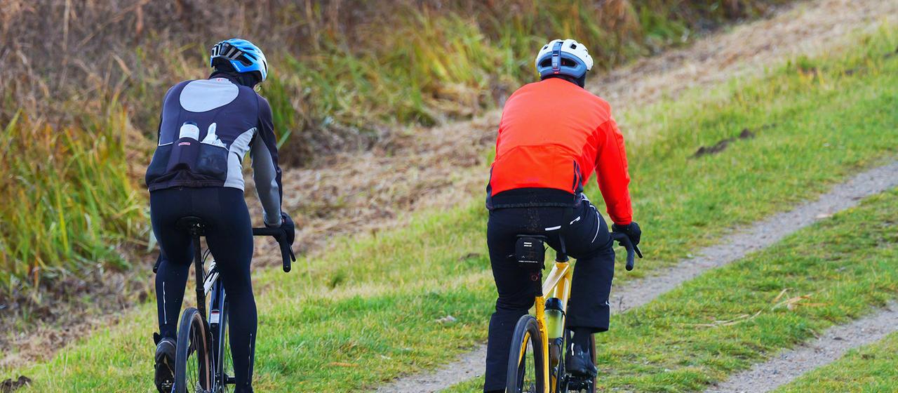 Two men in outdoor clothing ride gravel bikes along a dirt road. 