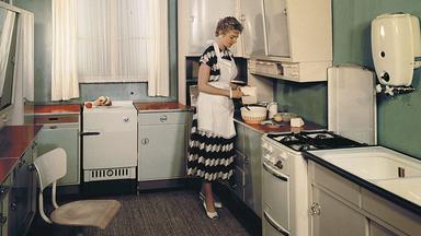 Eine Frau mit Schürze steht in einer 50er-Jahre Einbauküche und backt.