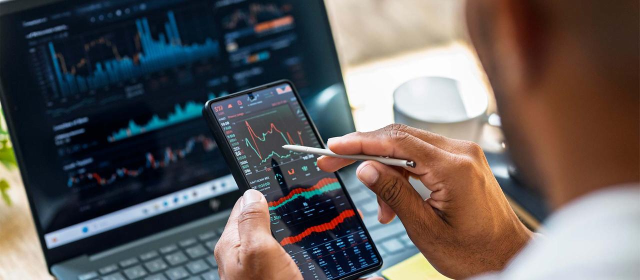 Eine Person überprüft Börsenkurse auf einem Smartphone- und Laptopbildschirm.