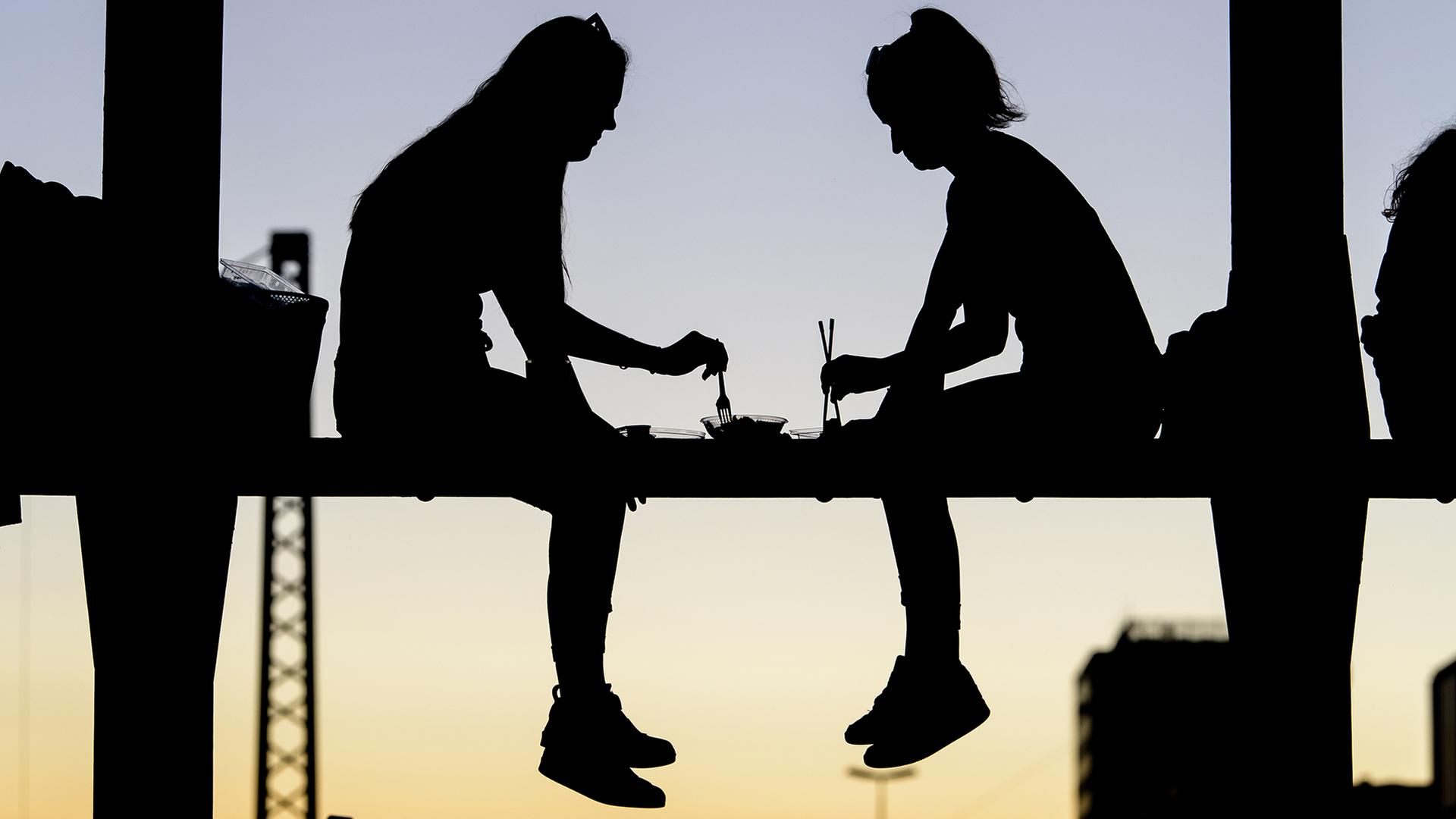 Zwei junge Frauen essen während des Sonnenuntergangs auf dem Geländer einer Brücke.