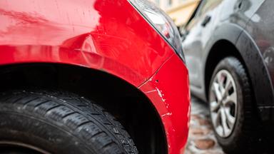 Ein rotes Autos steht mit einem Blechschaden in einer Parklücke, während ein graues Auto vorbei fährt.