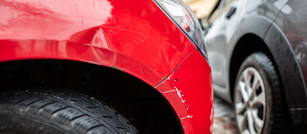 Ein rotes Autos steht mit einem Blechschaden in einer Parklücke, während ein graues Auto vorbei fährt.