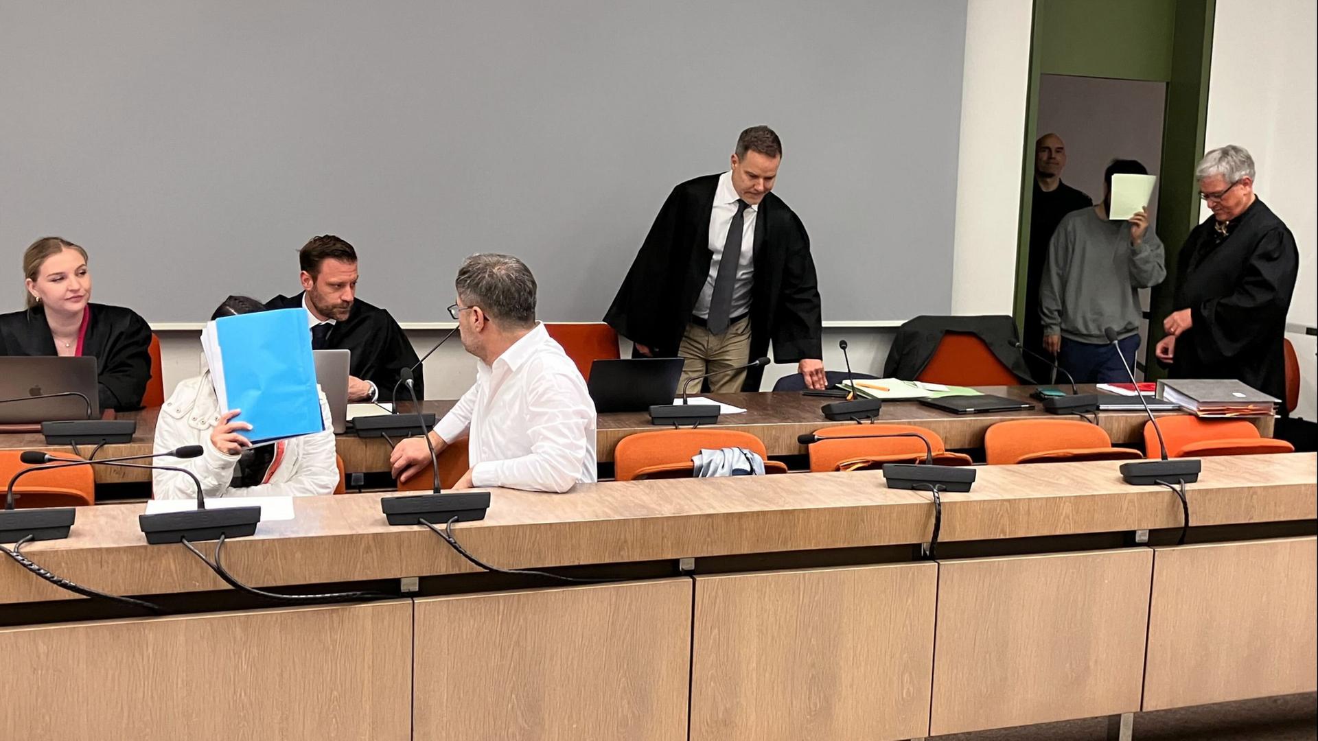 Die wegen Versklavung, Vergewaltigung und Verbrechen gegen die Menschlichkeit verurteilte irakische Frau (2.v.l mit Mappe vor dem Gesicht) sitzt im Saal des Oberlandesgerichts, während ihr Mann (hinten, 2.v.r) ebenfalls in den Saal kommt | Britta Schultejans/dpa