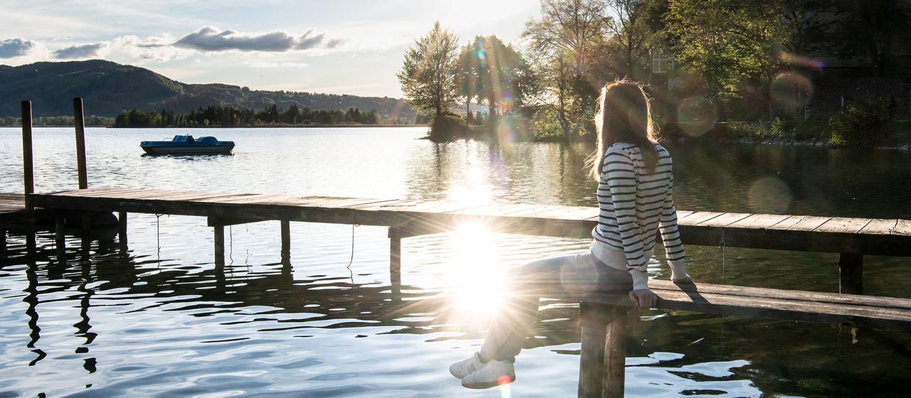 Eine Frau sitzt im Licht der Sonne auf einem Steg an einem See.