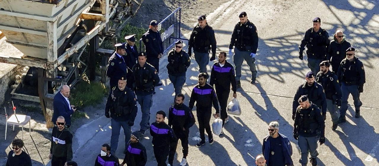 Migranten gehen im Hafen von Shengjin neben Sicherheitskräften, nachdem sie von einem Schiff der italienischen Marine im Nordwesten Albaniens an Land gegangen sind. (Archivbild vom 08.11.2024)