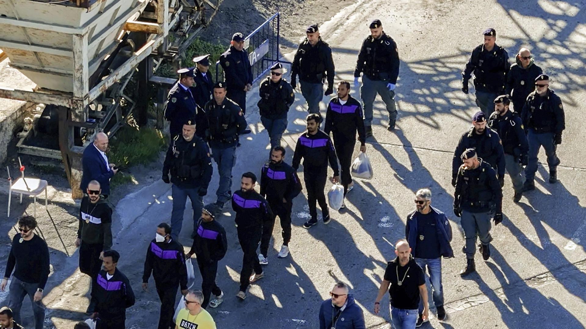 Migranten gehen im Hafen von Shengjin neben Sicherheitskräften, nachdem sie von einem Schiff der italienischen Marine im Nordwesten Albaniens an Land gegangen sind. (Archivbild vom 08.11.2024) | Vlasov Sulaj/AP/dpa