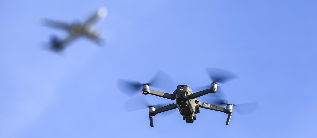 Eine Drohne fliegt am Himmel, im Hintergrund verschwommen ein Flugzeug.