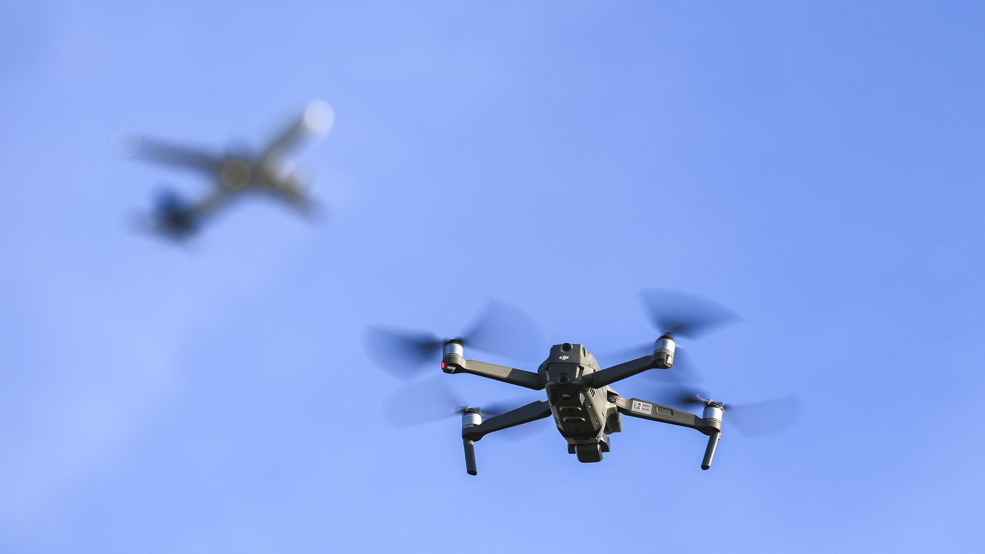 Eine Drohne fliegt am Himmel, im Hintergrund verschwommen ein Flugzeug. | dpa