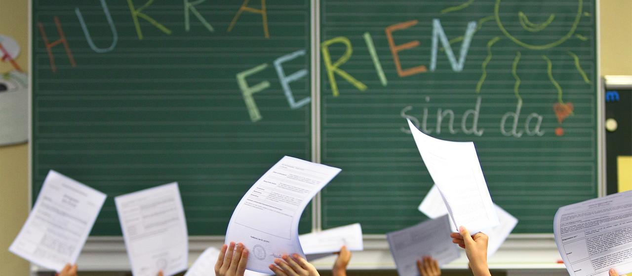 Schüler einer ersten Klasse halten ihre Zeugnisse vor einer Tafel mit der Aufschrift ''Hurra Ferien sind da''