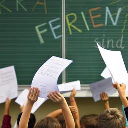 Schüler einer ersten Klasse halten ihre Zeugnisse vor einer Tafel mit der Aufschrift ''Hurra Ferien sind da''