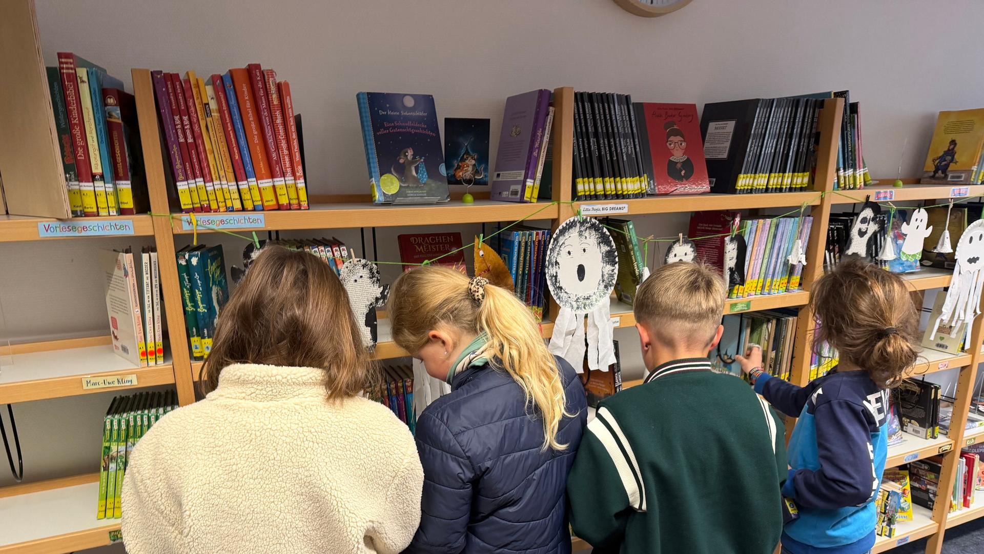 Vier Kinder tummeln sich vor einem Bücherregal und schauen alle interessiert das Sortiment an. An dem niedrigen Regal hängt Halloween-Deko mit kleinen Papier-Gespenstern. Zwei der Kinder blättern gerade gemeinsam in einem Buch. | Jörn Perske (hr)