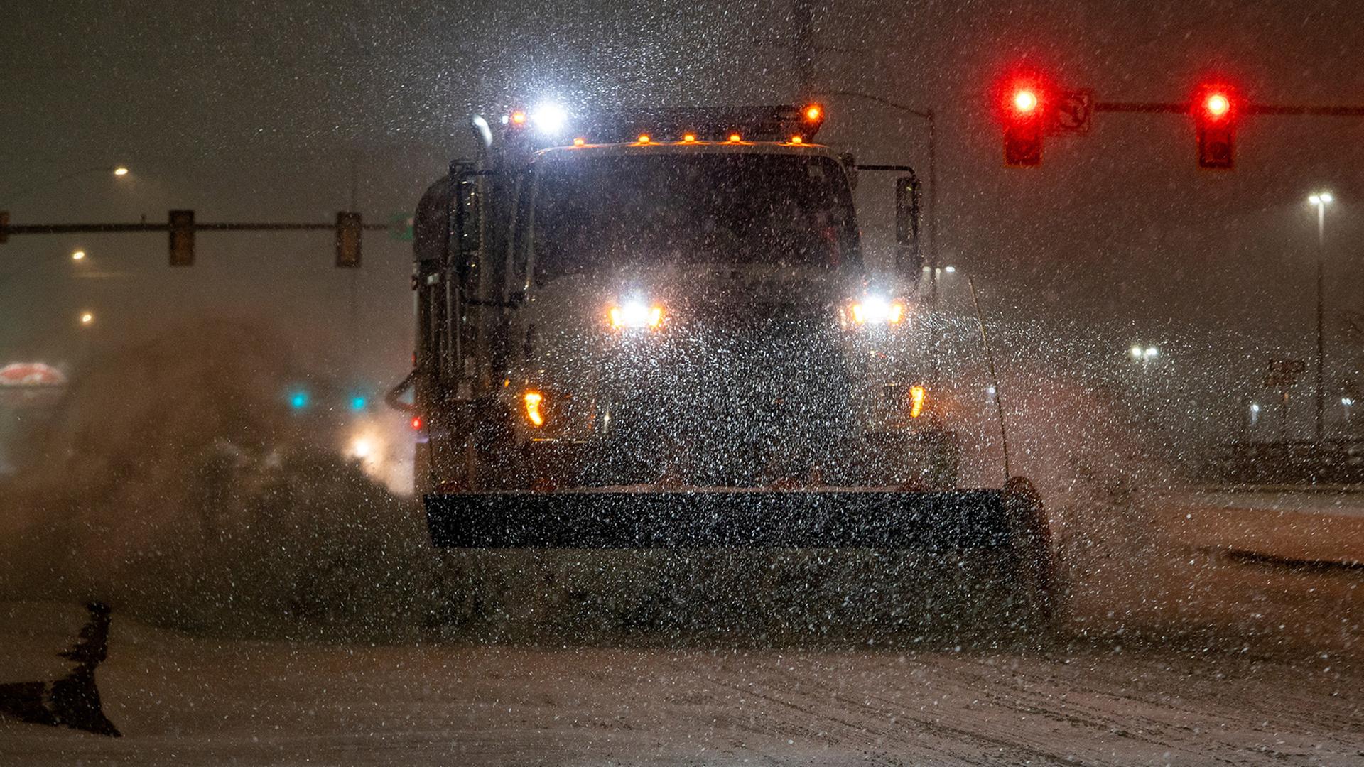 Ein Schneepflug fährt nachts durch starken Schneefall auf einer Straße, im Hintergrund sind rote Ampeln und Straßenlaternen zu sehen. | REUTERS