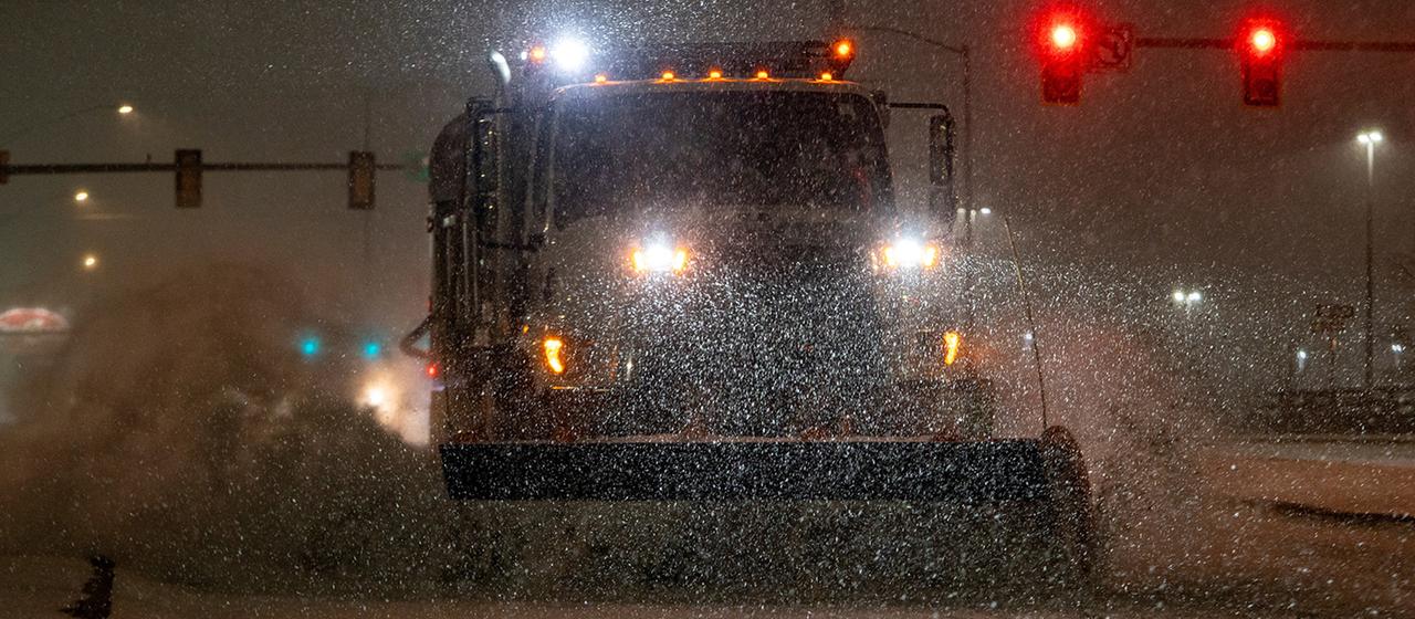 Ein Schneepflug fährt nachts durch starken Schneefall auf einer Straße, im Hintergrund sind rote Ampeln und Straßenlaternen zu sehen.