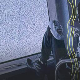 Ein Mann mit Maske sitzt auf einem Stuhl vor einem Fernseher. Auf seinem Knie liegt eine Maske mit Grimasse.