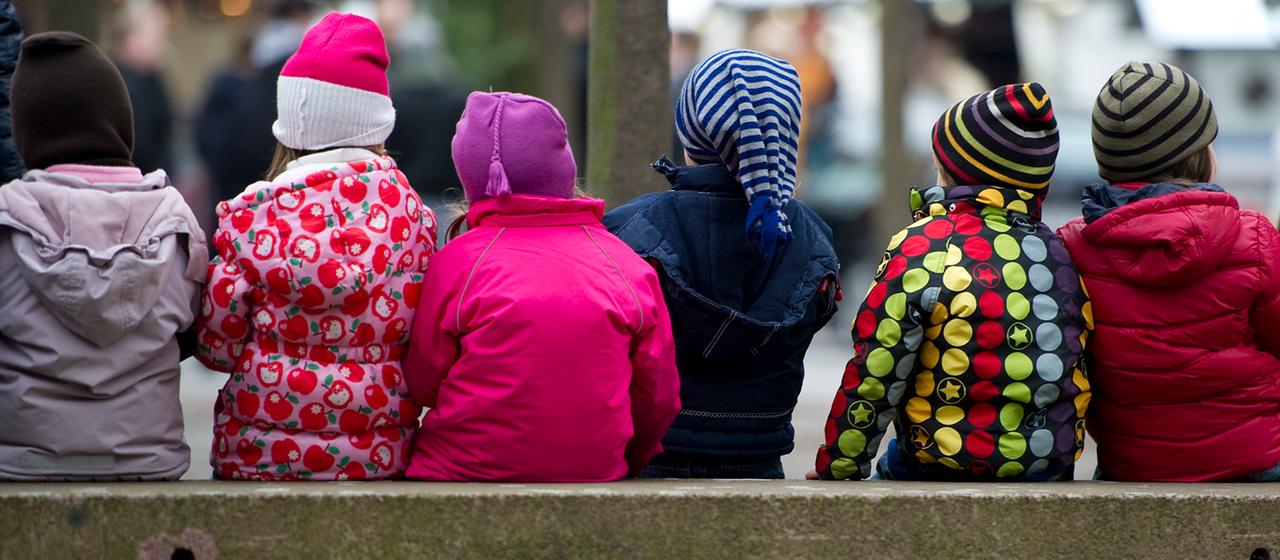 Kinder in Jacken und Mützen sitzen auf einer Bank.