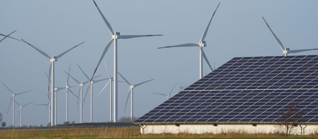Windenergieanlagen stehen neben einer Halle in Reußenköge (Schleswig-Holstein) mit Photovoltaik-Anlagen auf dem Dach.
