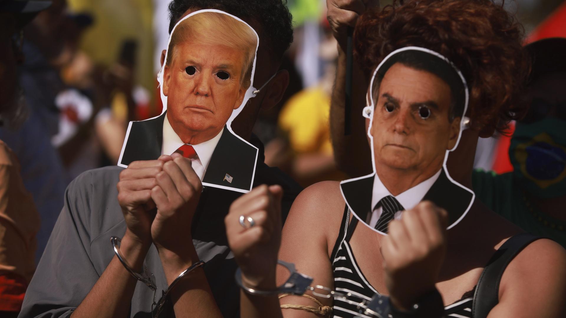 Demonstranten in symbolischen Handschellen halten Masken von US-Präsident Trump und dem ehemaligen brasilianischen Präsidenten Bolsonaro hoch. | dpa