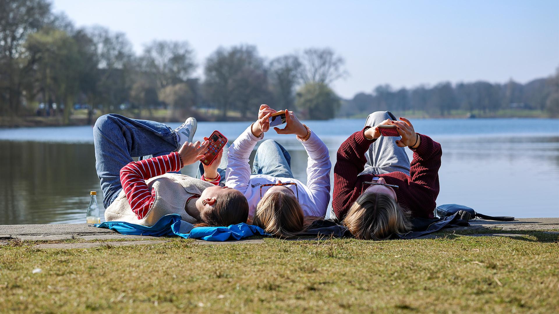 Drei junge Frauen liegen am Ufer eines Sees in der Sonne.