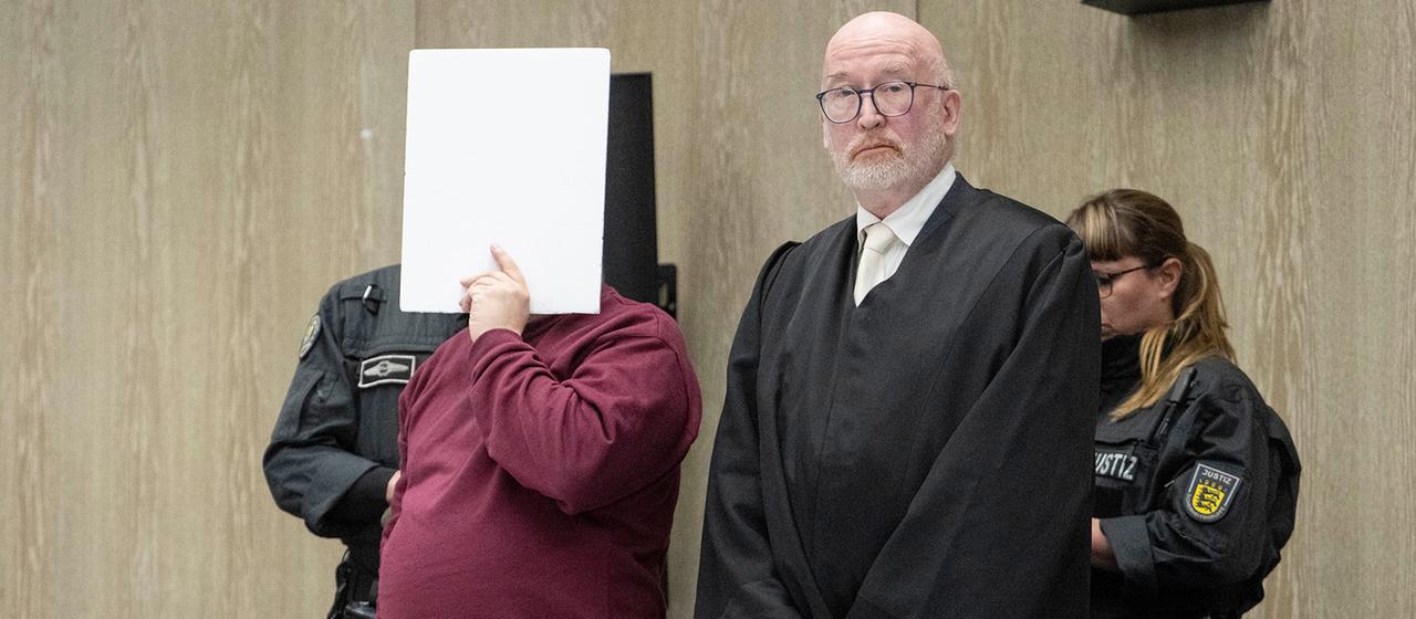 Der Angeklagte steht mit einem Aktendeckel vor seinem Gesicht im Gerichtssaal.