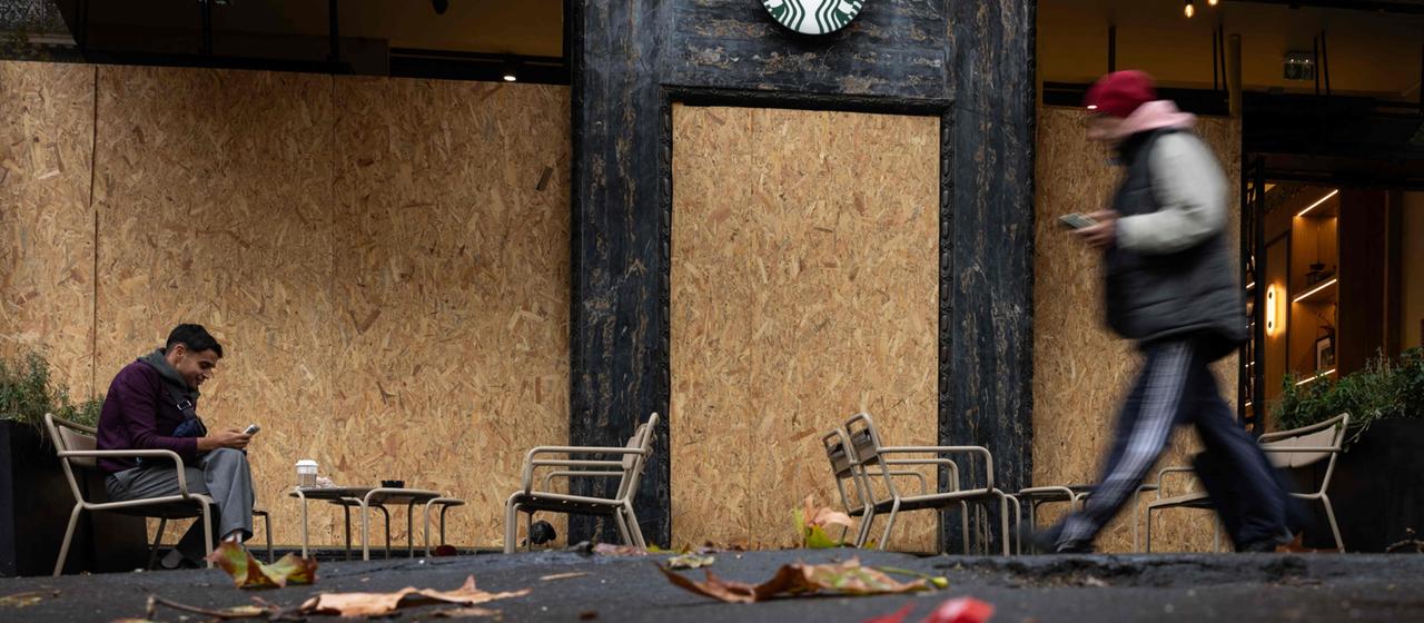 Ein Mann sitzt mit einem Getränk auf der Terrasse eines Starbucks-Cafés in Paris, dessen Fenster mit Sperrholzplatten vernagelt sind.