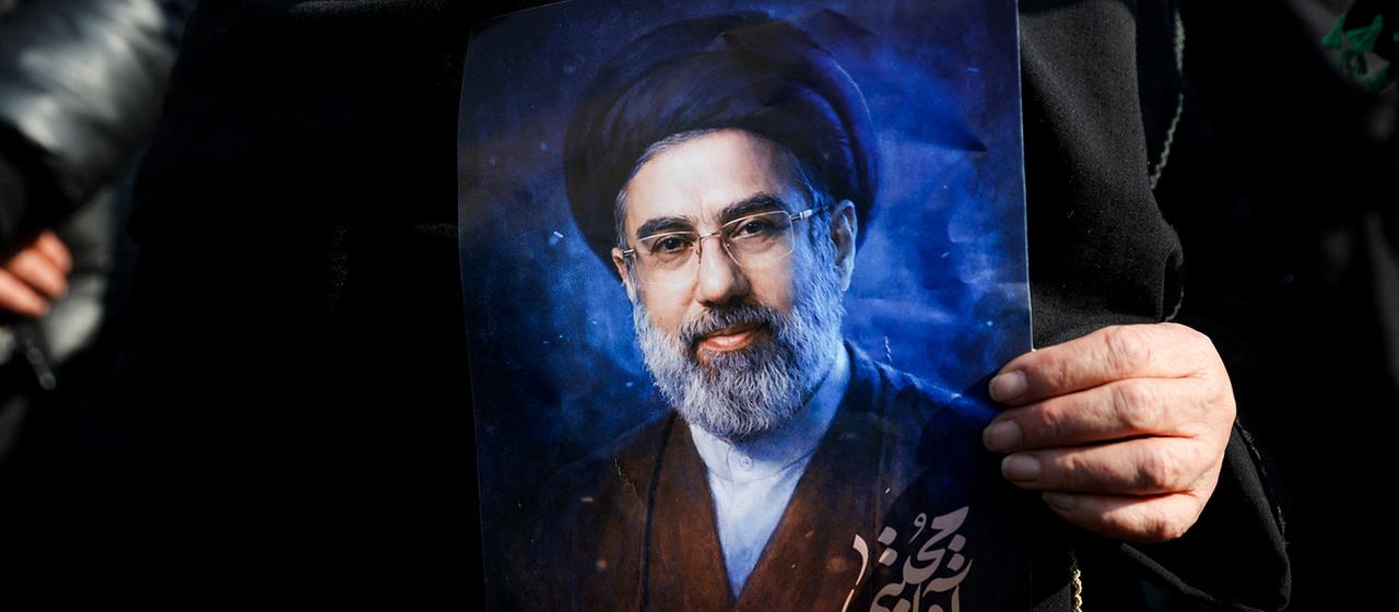 A woman holds a picture of Modshtaba Khamenei in her hands.